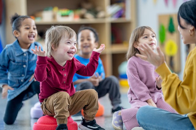 A small group of preschool children sit on the floor in front f their teacher as she leads them in a...