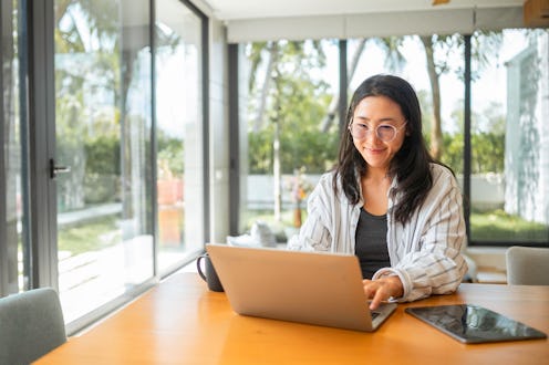An Asian woman exudes joy as she works from home, comfortably seated with her laptop and digital tab...