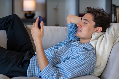 Young man is lying comfortably on a sofa, using his smartphone with a relaxed expression, enjoying h...