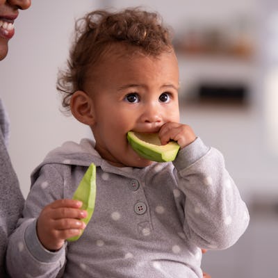A baby of about 9 months eats an avocado, holding a half of the fruit in each hand.