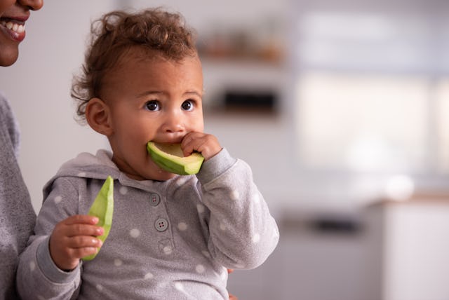 A baby of about 9 months eats an avocado, holding a half of the fruit in each hand.
