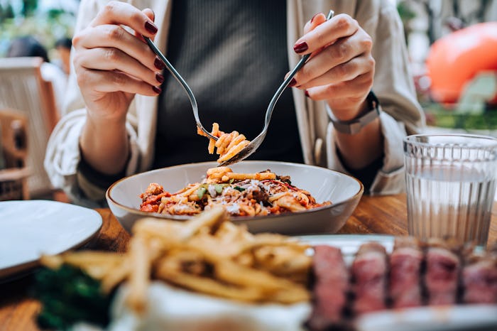 Close-up, mid-section of young Asian woman sitting at dining table holding cutleries, twirling fresh…