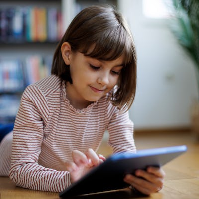A child enters something into her tablet while lying on the floor of her home.