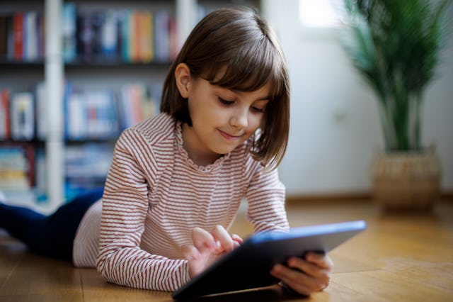 A child enters something into her tablet while lying on the floor of her home.