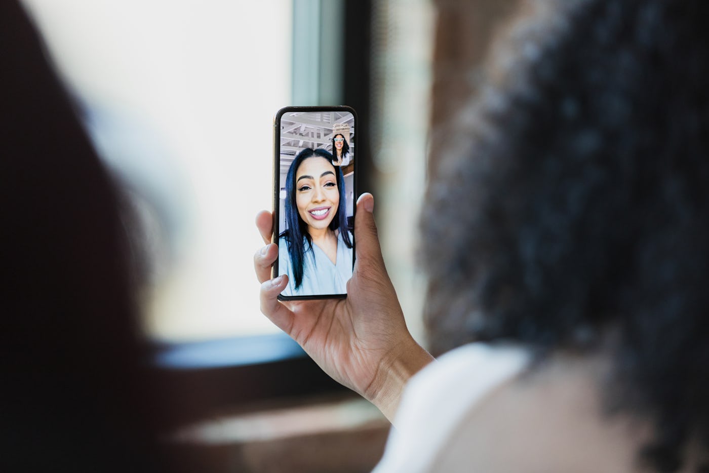 Two unrecognizable women use a teleconferencing app on a smart phone to meet with a female colleague.