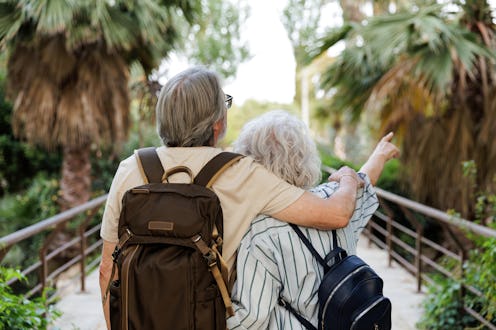 Elderly couple with backpacks enjoying a leisurely walk through a lush tropical garden, embracing th...