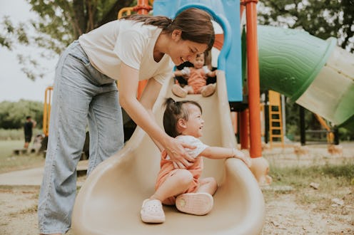 Smiling mother is embracing toddler while playing sliding equipment at the playground.