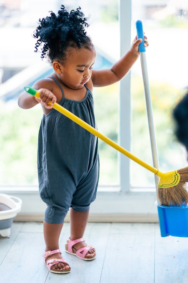 A toddler with curly hair sweeps the floor of her home. She is wearing a denim romper and pink sanda...