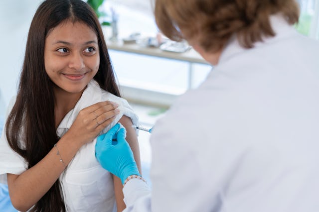 A teenage girl with long brown hair smiles as a doctor, with her back to the viewer, administers a v...