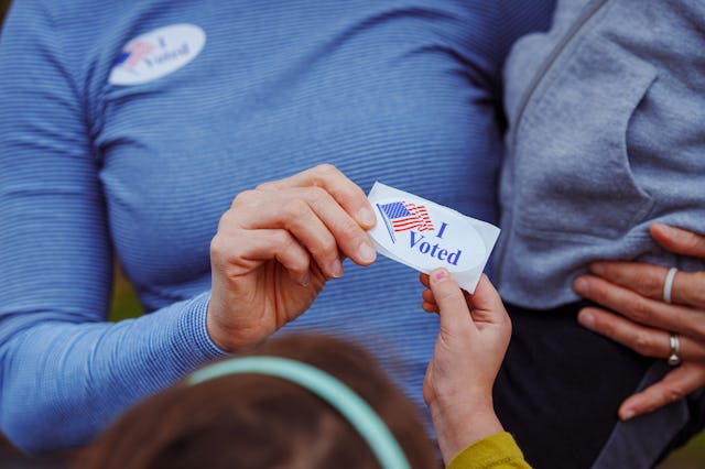 Cropped view of an unrecognizable woman holding her toddler on her hip and handing a sticker that sa...