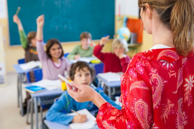 Students raise their hand as a teacher in the foreground, with her back to the viewer, calls on one ...