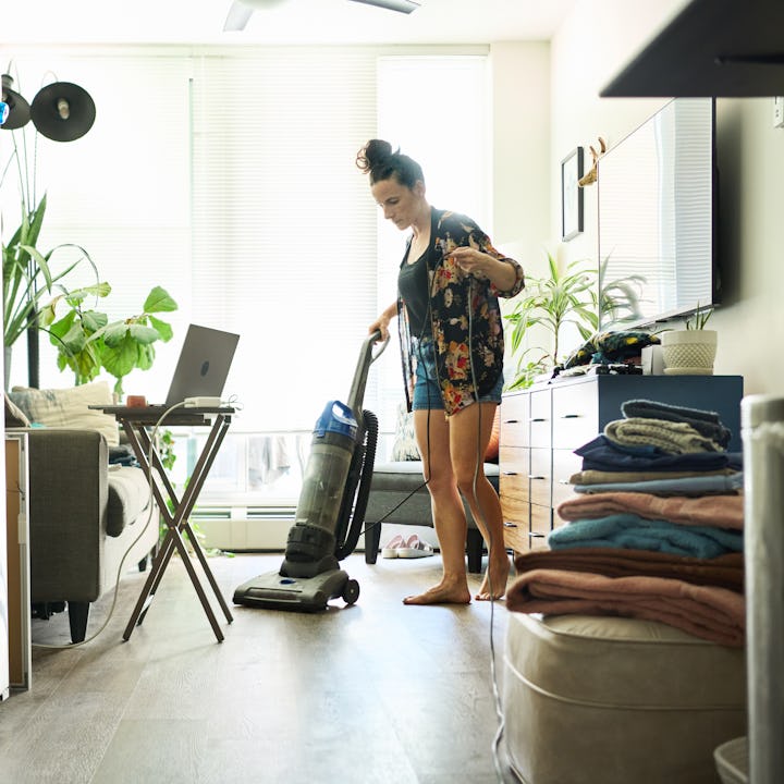 Young woman vacuuming her living room floor while doing laundry and chores around the home at the we...