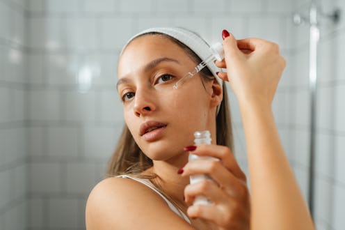 Young woman standing in a bright, modern bathroom, applying facial serum to her skin. She holds a sm...