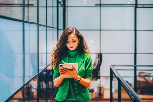 Businesswoman dressed in a vibrant green blazer walking in a contemporary office space while working...