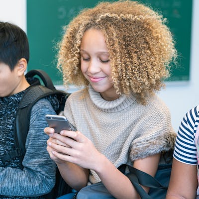 A group of middle school students is sitting in a row on the desks in their classroom. All of the students are using their mobile phones.