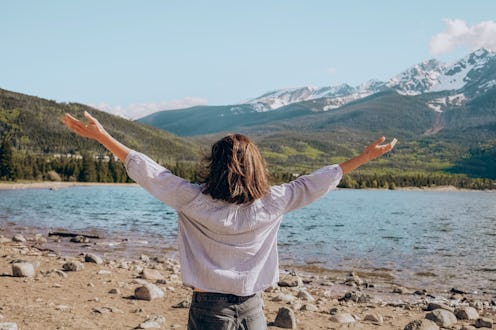 woman stands on a rocky shore, arms outstretched toward a mountain lake. The background features lus...