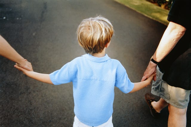 A young child wearing a light blue shirt walks on a path, holding hands with adults on either side, ...