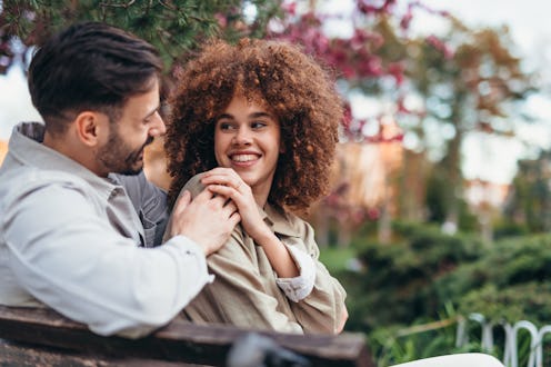 Young couple sitting on a park bench, enjoying each other's company while sharing affectionate smile...