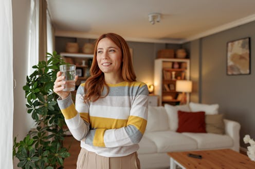 Young woman holding a glass of water, gazing out the window and smiling, savoring a peaceful moment ...