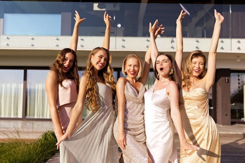 Portrait of excited young women wearing elegant dresses standing in front of villa and raising hands...