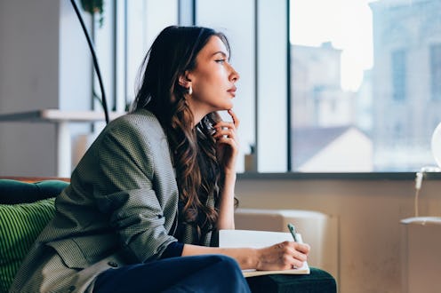 A professional woman sits pensively holding a notebook and pen, working in a contemporary office env...
