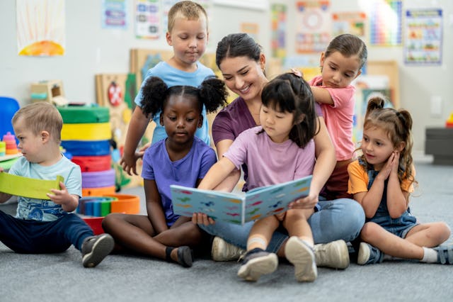 A preschool teacher sits with a small group of children as she reads them a story. They are dressed...