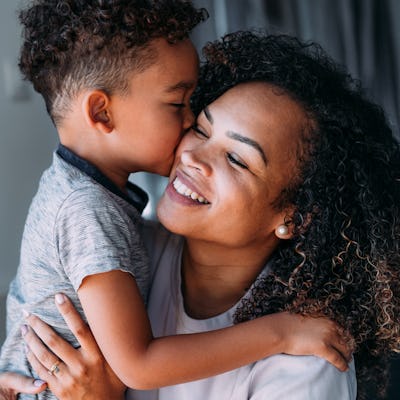 A joyful mother and her young son share a loving moment, with the boy giving her a kiss on the cheek. They both smile, surrounded by soft lighting.