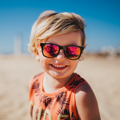 Close up of a little blonde boy wearing sunglasses on the beach smiling to the camera, in a story listing out cool boy baby names.