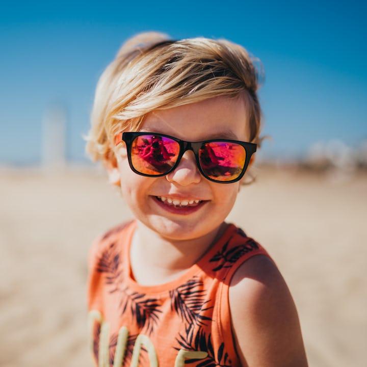 Close up of a little blonde boy wearing sunglasses on the beach smiling to the camera, in a story li...