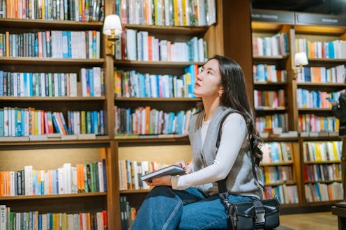 Young Asian woman sitting on bench looking for books from bookshelf in book store.