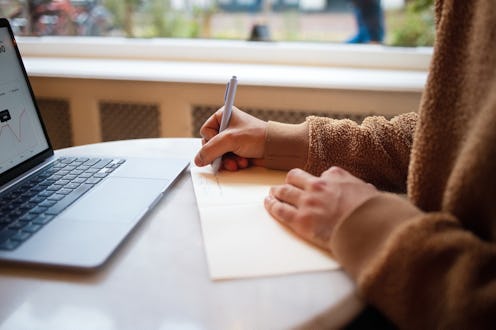 A photograph of a person writing in a notebook, seated beside a laptop in a cozy cafe, with focus on...