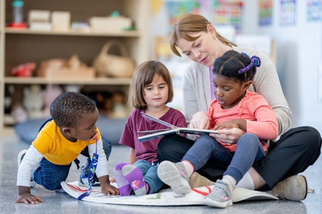 A daycare teacher sits on the floor of the classroom with three young children as she reads a story ...