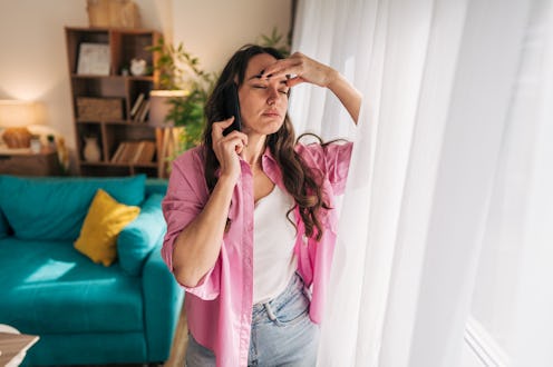 Young woman standing by the window, holding her head with one hand while talking on the phone, displ...