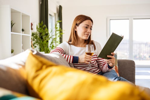 Happy woman holding glass of white wine on sofa reading book