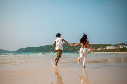 A Hispanic young woman and a Black man, both in casual beachwear, run hand in hand along a sunlit tr...