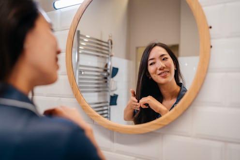 A young woman is styling her hair in a modern bathroom, using a round brush and a hairdryer. Dressed...