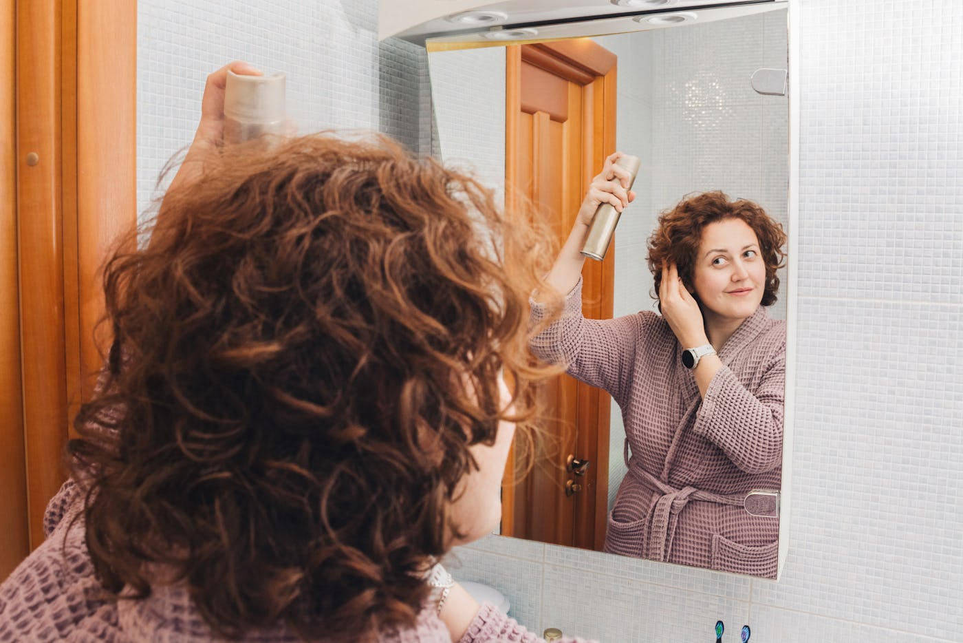 Woman with curly hair in bathrobe stands in front of mirror in bathroom and using hair spray for styling hair. Use of hair cosmetics, home self-care. Routine process of self-care, importance of communication and Home beauty rituals. Copy space