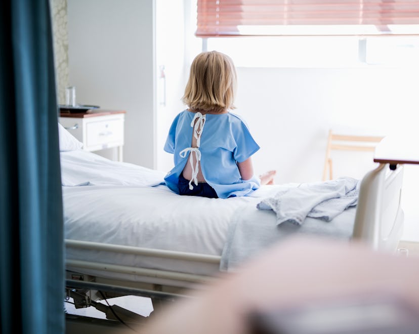 Rear view of ill girl sitting on bed. Female child patient is wearing hospital gown. She is resting ...