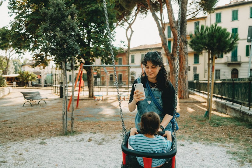 Mother is pushing her son on a swing while looking at her phone on a sunny day at the playground