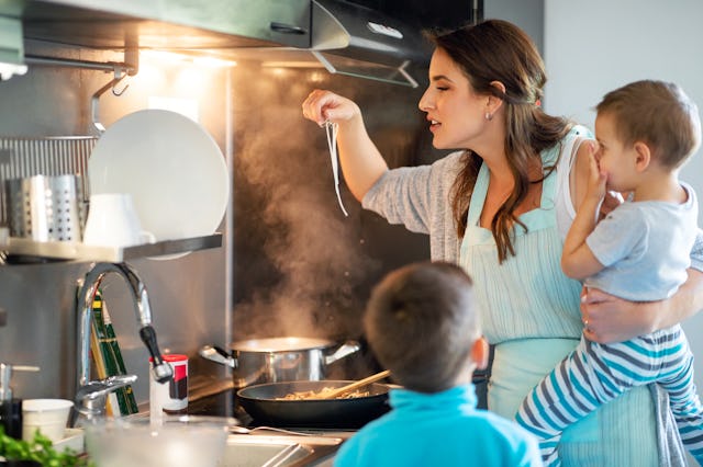 Happy mother tasting spaghetti by the stove and carrying toddler on hip, older son standing by them