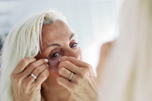 Attractive mature woman looks in mirror and applying on her face hydrogel under-eye recovery patches...