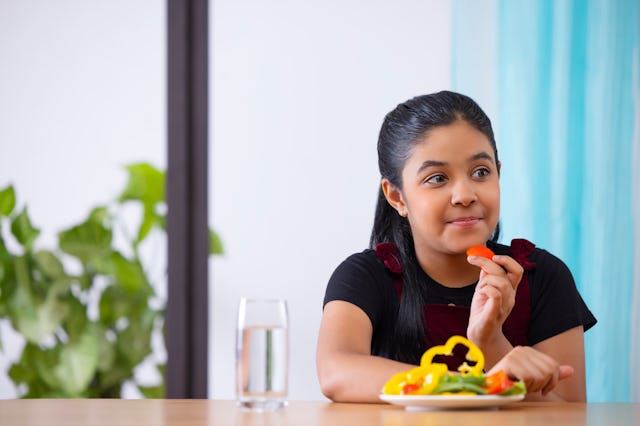 A young girl sitting at a table enjoys a piece of food, with a colorful plate of salad in front of h...