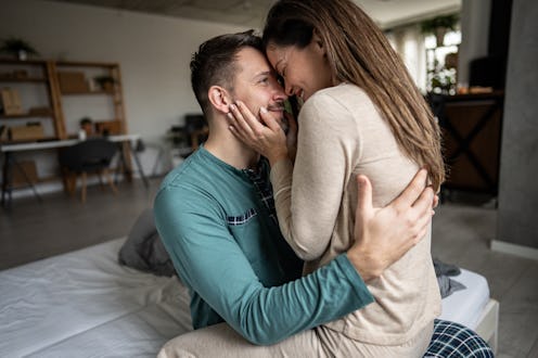 Young couple embracing and smiling while sitting on a bed at home, sharing moments of intimacy and j...