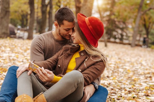 Romantic young couple sitting on fallen autumn leaves in city park and taking selfie on mobile phone