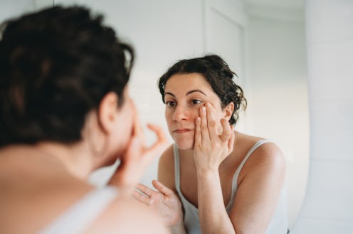Young woman examining her reflection in the mirror, gently touching her skin while checking for impe...