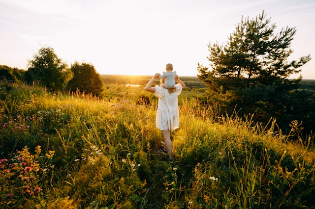 Mother carrying baby on shoulders while walking in meadow at sunset