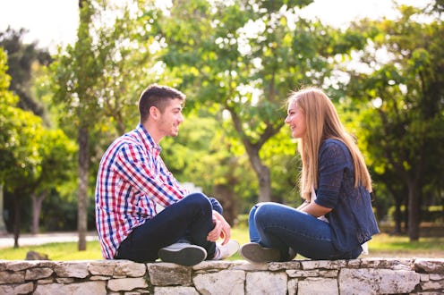 Two young adults sit on a stone wall in a park, engaged in an animated conversation. Sunlight filter...