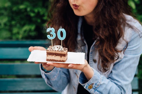 A cheerful woman holds a delicious chocolate birthday cake decorated with lit candles forming the nu...