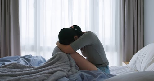 asian woman sitting on a bed hugging a blanket and looking distressed