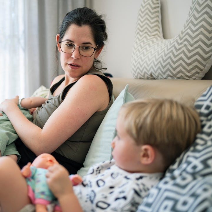 Mother breastfeeding her baby on a sofa at home looking at her toddler son plays with a doll in the ...